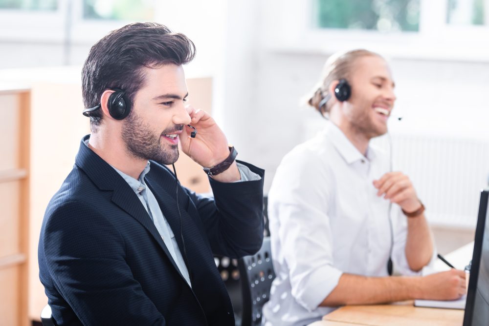 Two men wearing headsets sit at desks in an office. One man is talking, while the other smiles and laughs using a computer as they discuss voluntary benefits with clients.