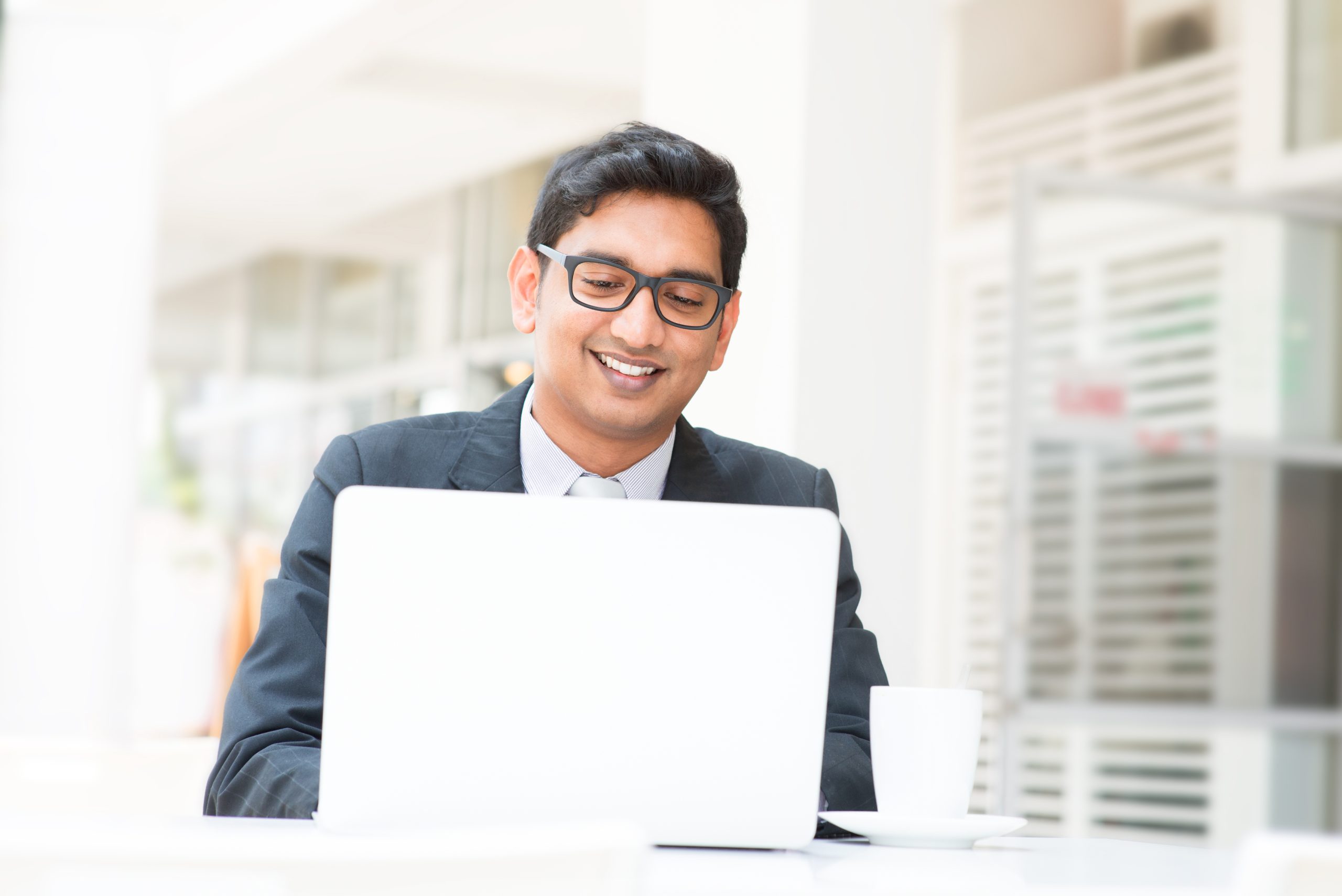 A man wearing glasses and a suit is smiling while working on a laptop at a table with a white coffee cup, representing the professional spirit of Great Choice Agency.