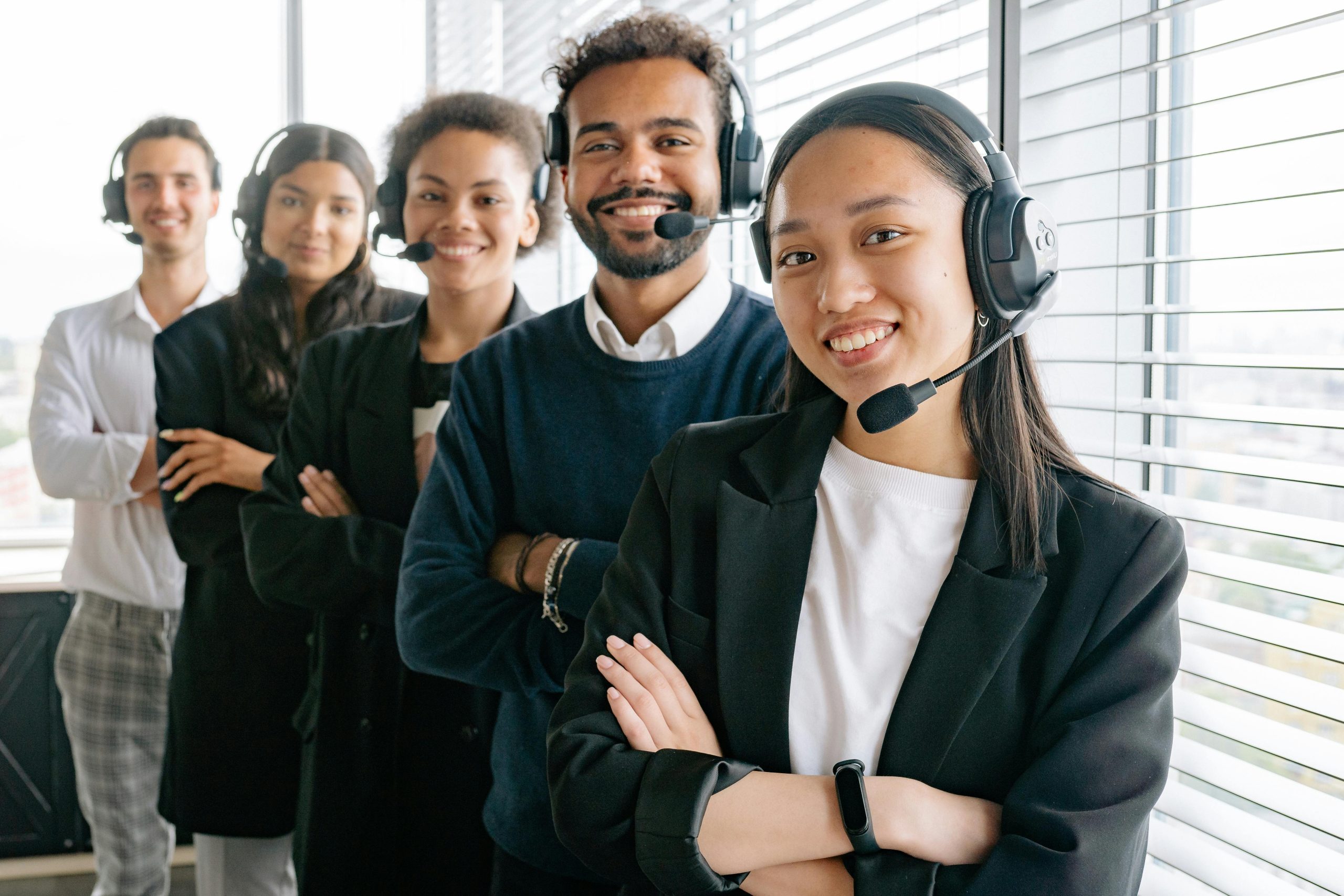 Five diverse customer service representatives wearing headsets stand in a line with arms crossed, smiling, in a modern office setting near large windows.