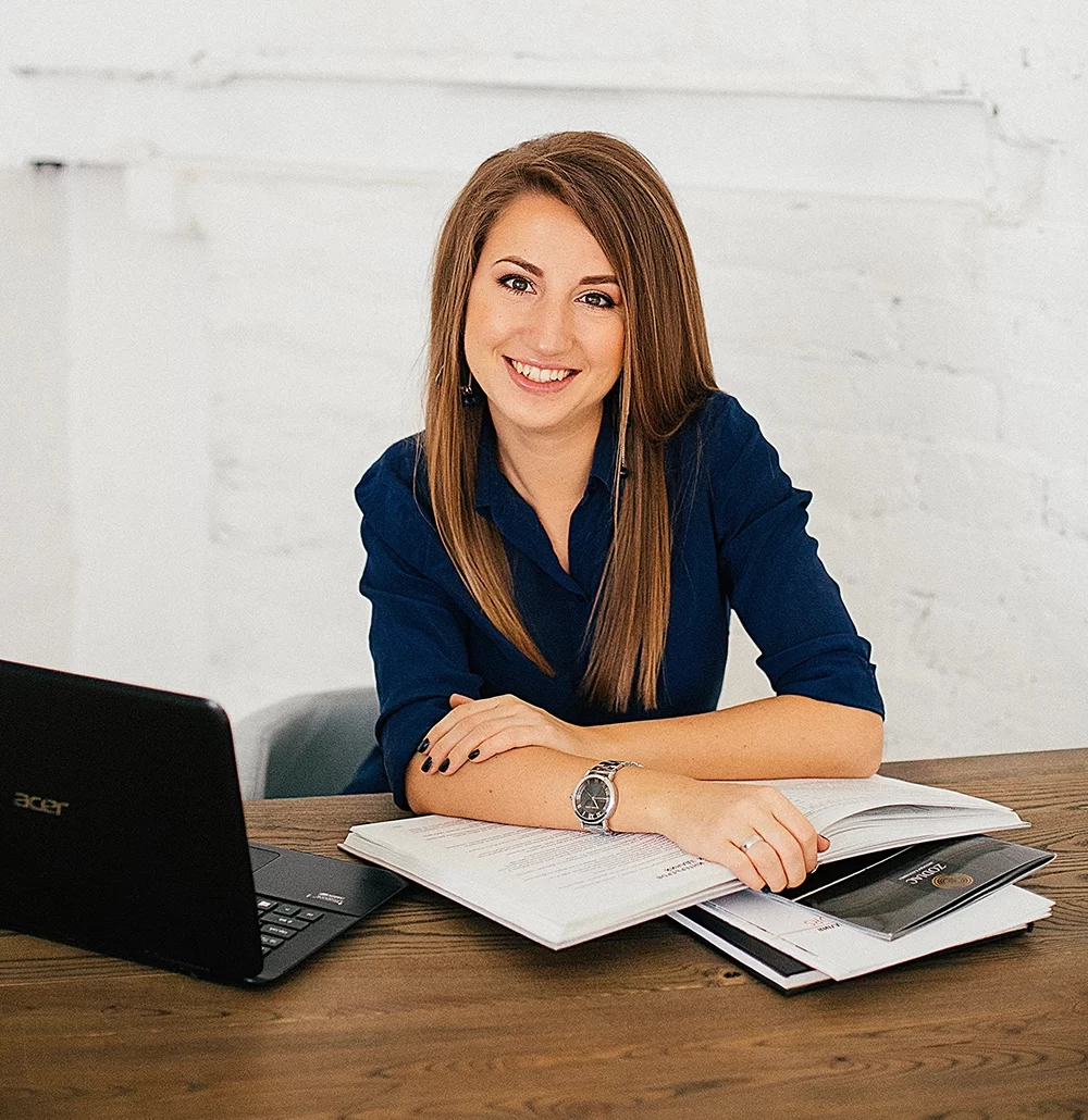 A woman sits at a desk with a laptop, notebook, and documents labeled "Voluntary Benefits," smiling at the camera in a bright, minimalist office setting.